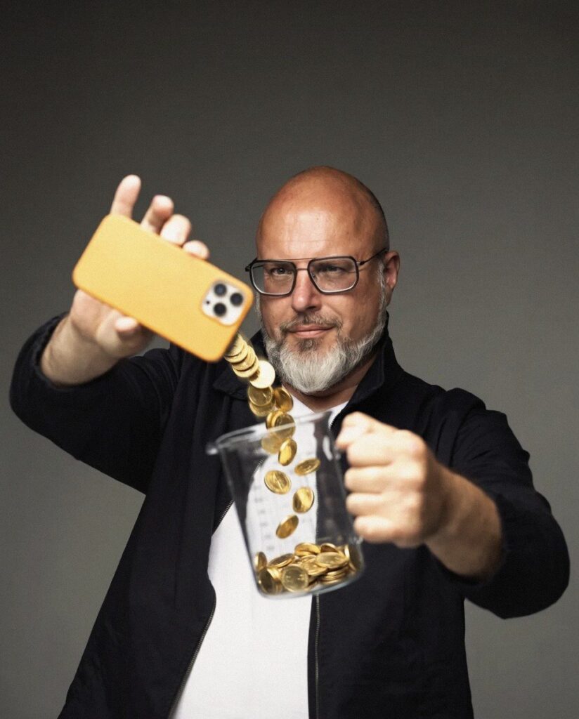 Sascha Schulz holds an orange smartphone from which gold coins pour into a glass measuring jug